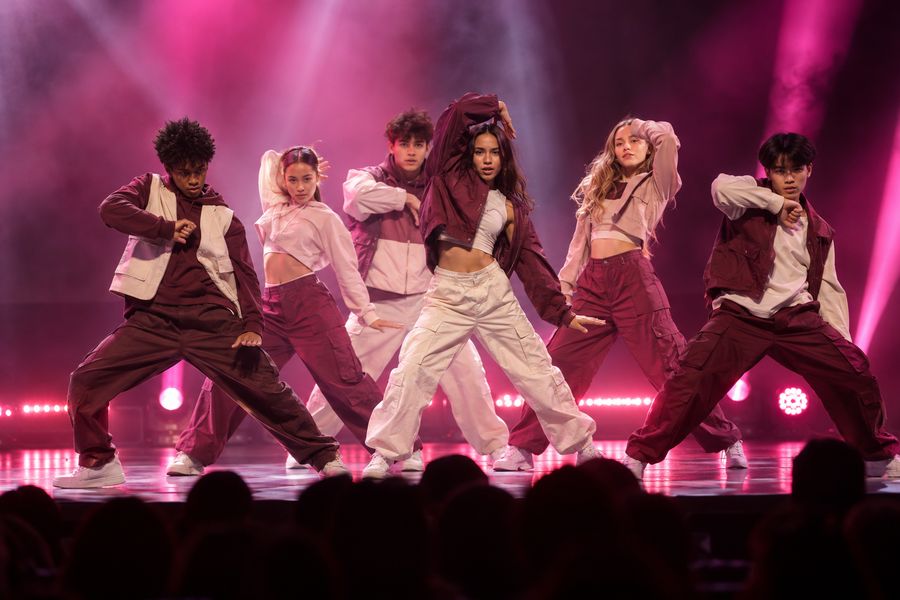 A teen hip-hop crew of boys and girls hitting a pose on a competition stage under pink lights