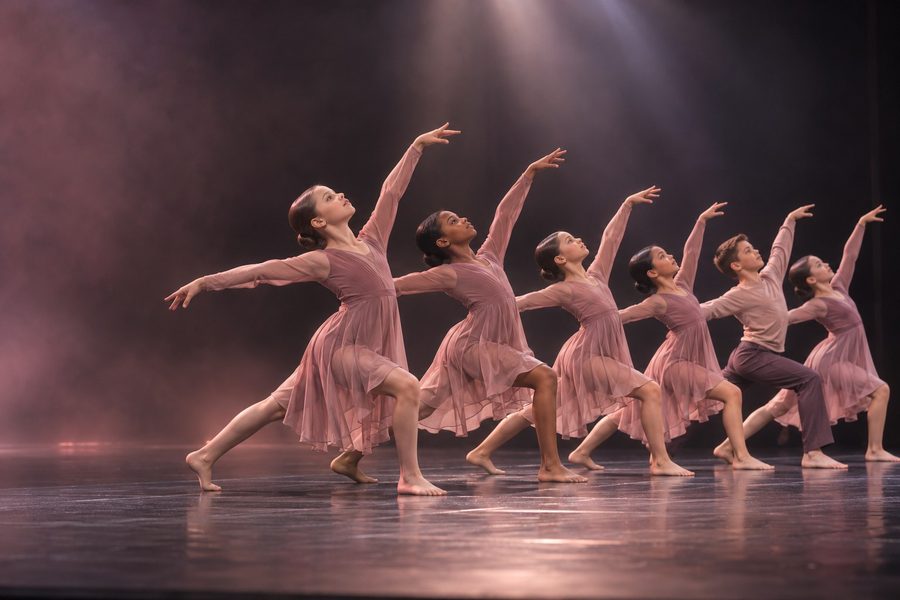 Six young contemporary dancers in matching blush dresses and identical buns, moving in unison on a competition stage